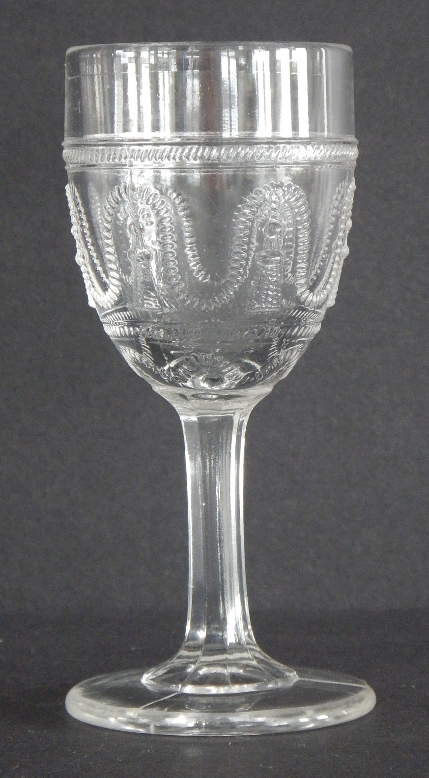 Close up of an antique clear glass cordial, Cord and Tassel pattern on the sides.